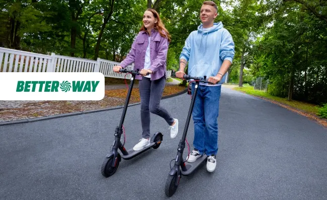 Dos personas en patinetes negros sobre asfalto gris, rodeadas de árboles y una valla blanca.