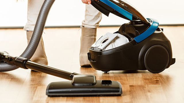 Persona aspirando un piso de madera con una aspiradora negra con detalles azules y tubo gris.