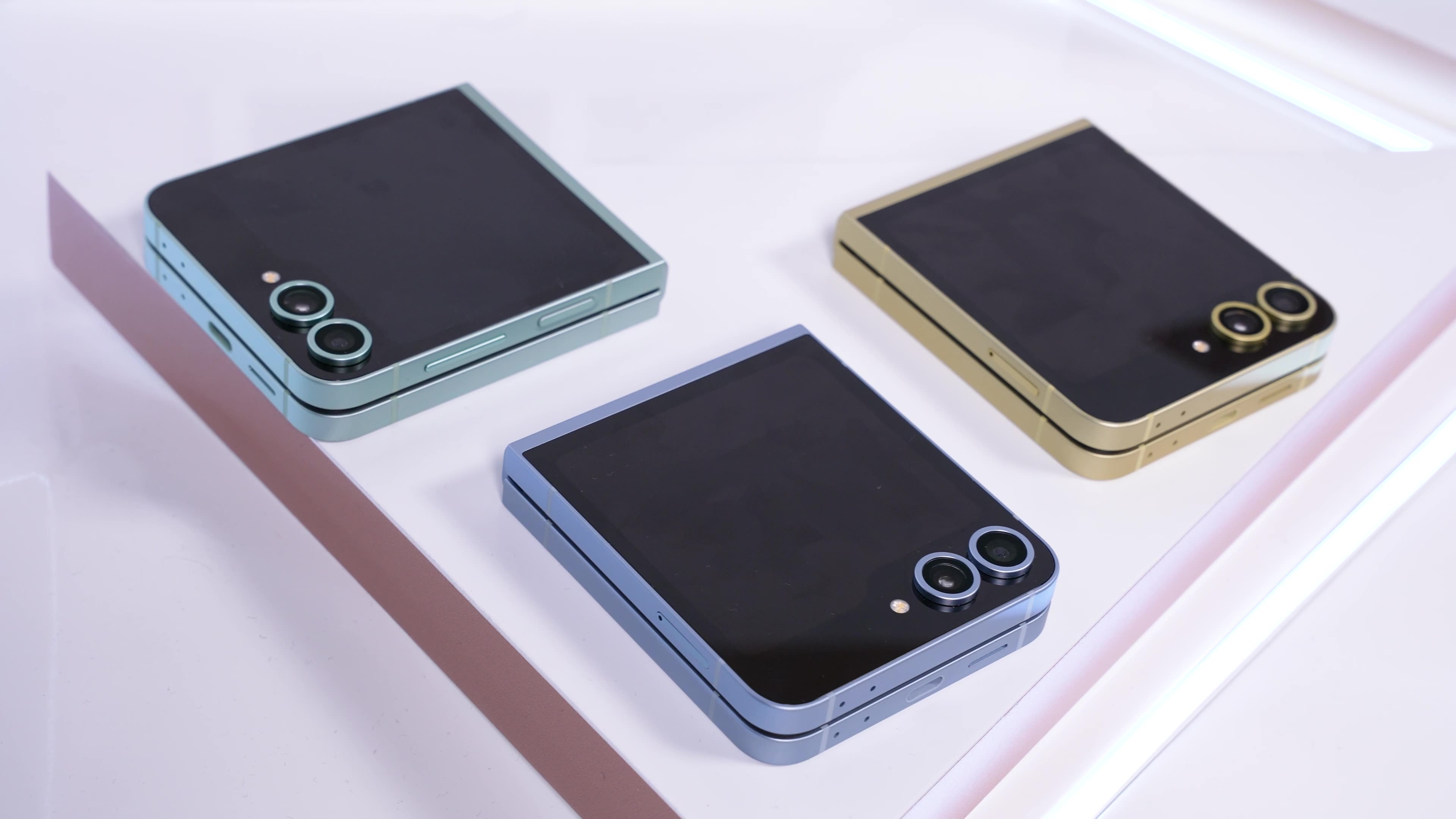 Tres teléfonos plegables cerrados, con marcos en azul, verde y dorado, sobre una superficie blanca con detalles lineales rosados.