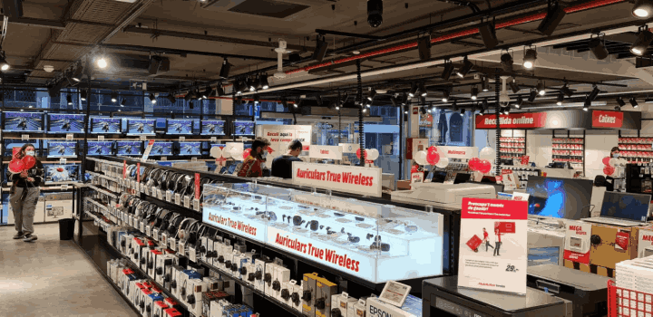 Tienda con pasillos llenos de productos electrónicos como auriculares, televisores y ordenadores. Luces en el techo.