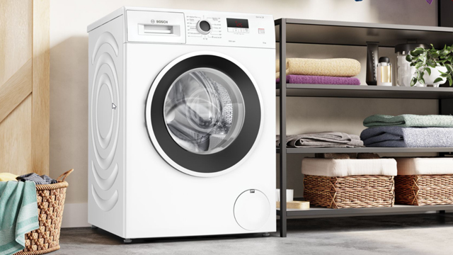 Lavadora de libre instalaci´n blanca, instalada en un amplio cuarto de lavado, junto a una estantería con ropa limpia y un cesto de ropa para lavar