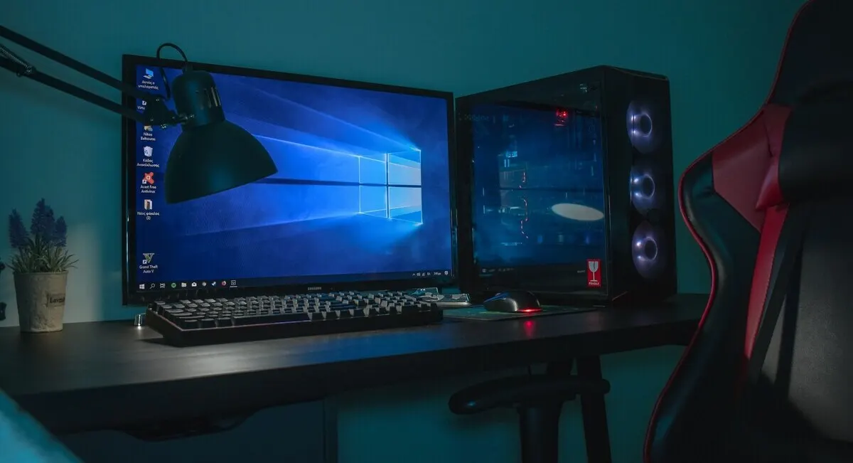 Setup gamer con PC iluminado, teclado mecánico, monitor con Windows 10 y silla ergonómica roja y negra.