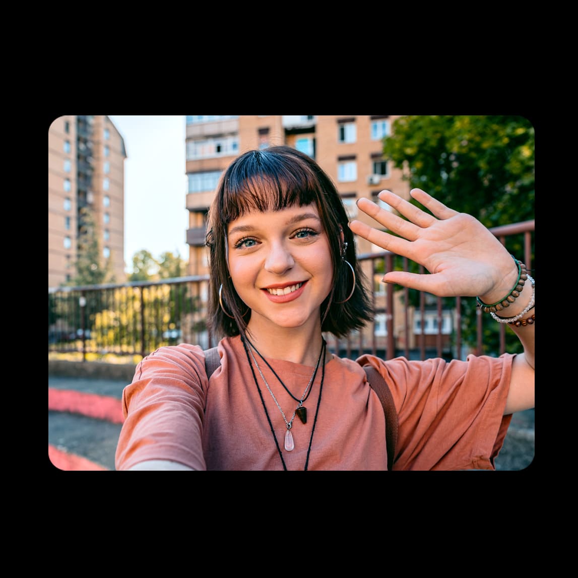 Photographie prise avec un smartphone Samsung montrant le portrait d'une jeune femme souriante et faisant un signe de la main dans un environnement urbain extérieur.