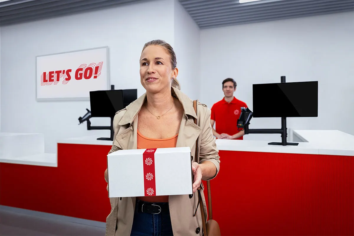 Mujer con caja blanca y cinta roja en tienda con mostrador rojo y blanco. Empleado con camisa roja al fondo y letrero "Let's Go!".