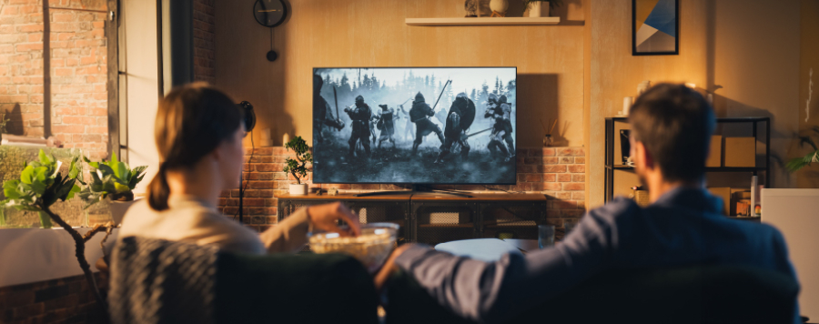 Una pareja sentada en un sofá viendo una película de guerra en la televisión. Hay palomitas y plantas en la sala de estar.