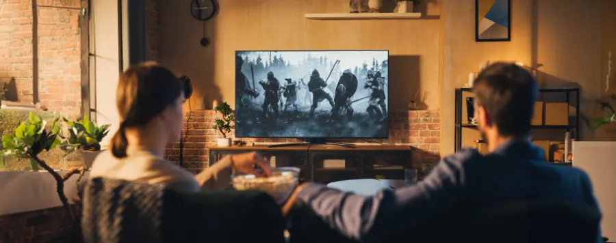 Una pareja sentada en un sofá viendo una película de guerra en la televisión. Hay palomitas y plantas en la sala de estar.