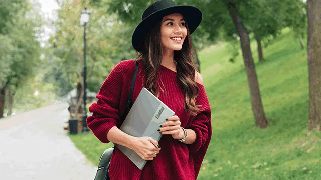 Mujer sonriente con sombrero negro y suéter rojo sostiene una laptop gris en un parque con árboles y una farola.