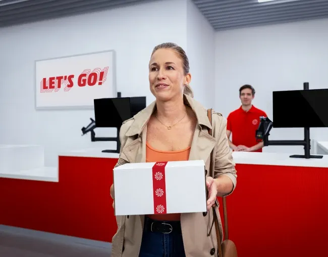 Mujer con caja blanca y cinta roja en tienda roja y blanca, con empleado al fondo y letrero "Let's Go!".