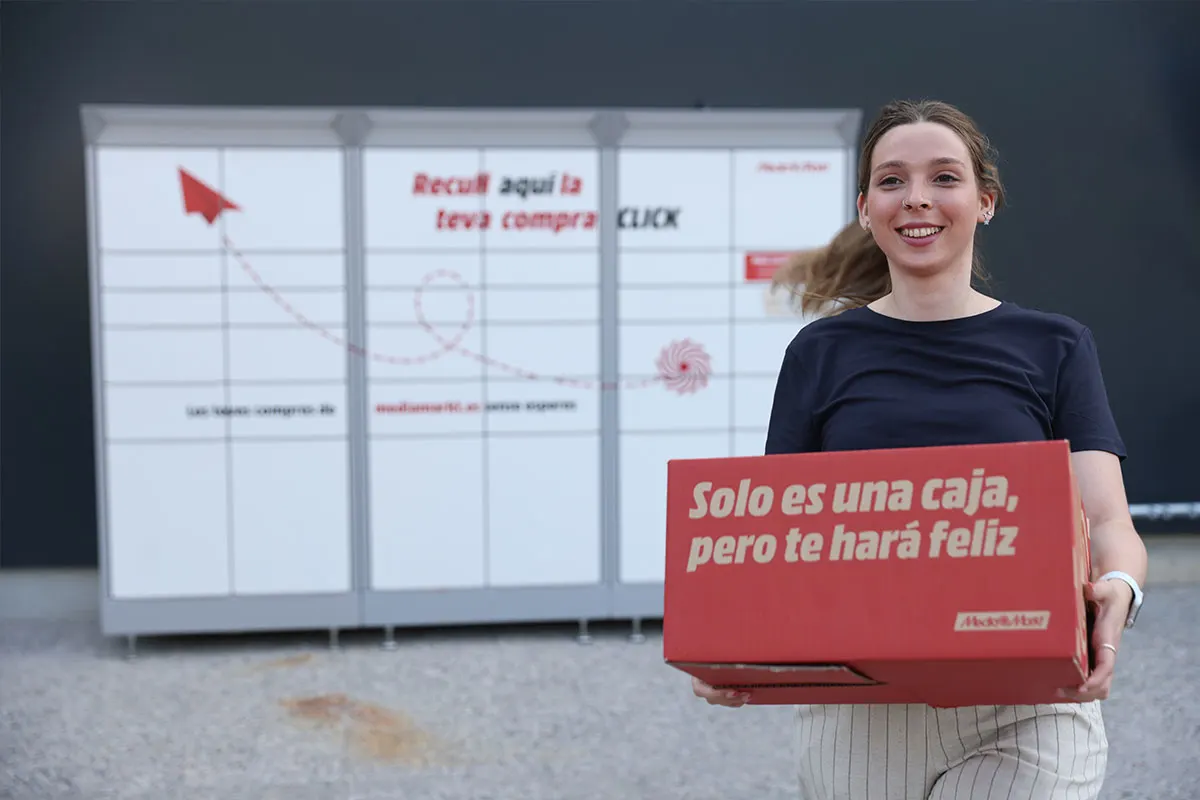 Una mujer sonriente sostiene una caja roja con texto frente a una taquilla blanca con gráficos e instrucciones.