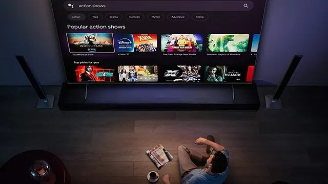 Persona viendo un televisor grande con "action shows" en la pantalla, con libro y taza en el suelo.