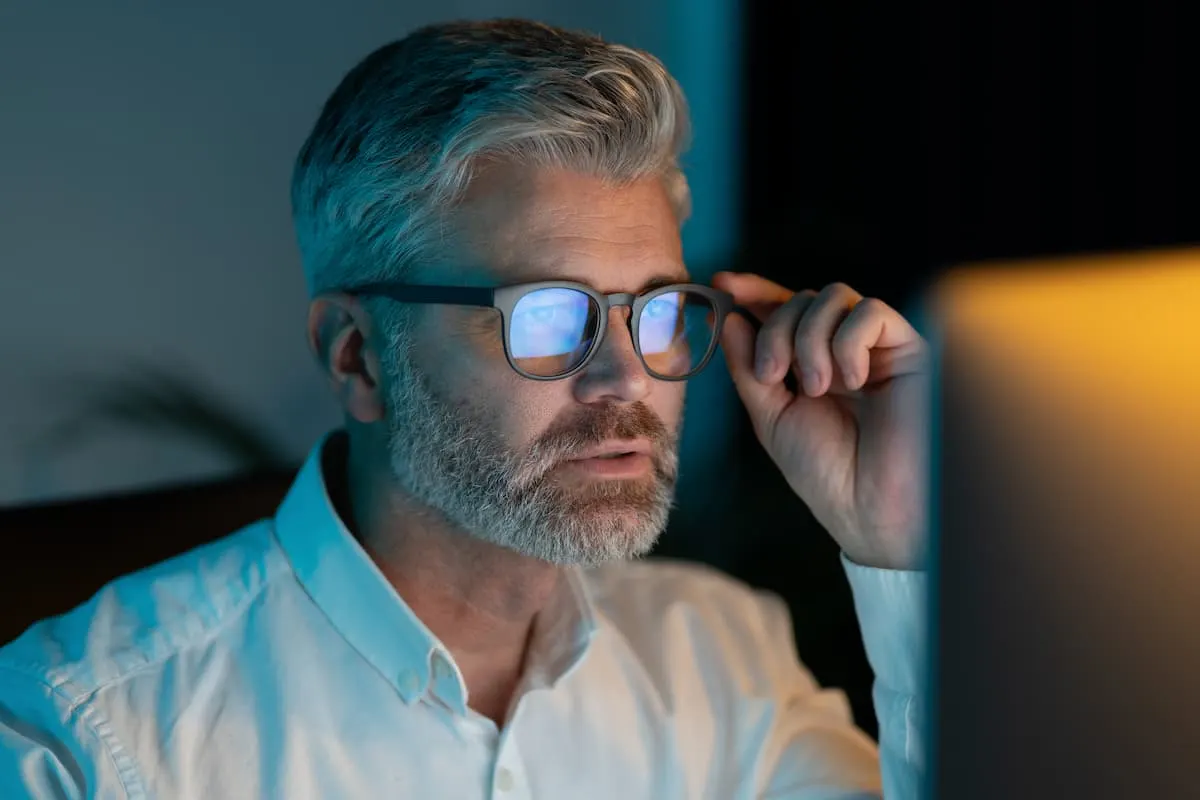 Hombre con gafas mirando la pantalla de un ordenador en una habitación poco iluminada.