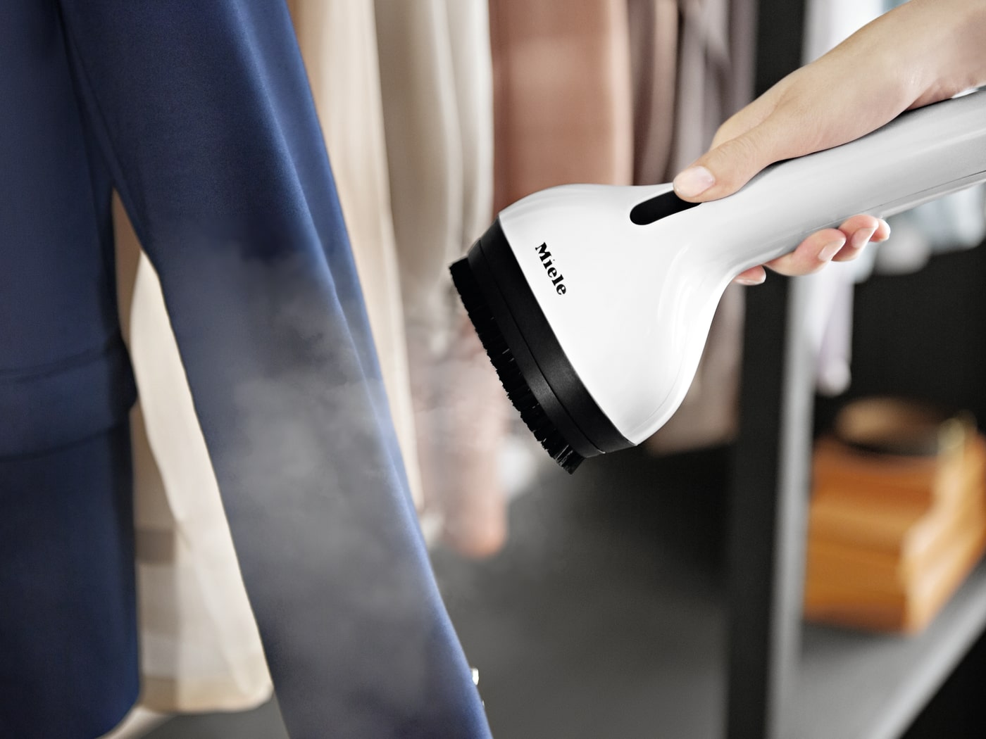 Plancha vertical blanca Miele vaporizando una prenda azul en un armario con ropa colgada al fondo.