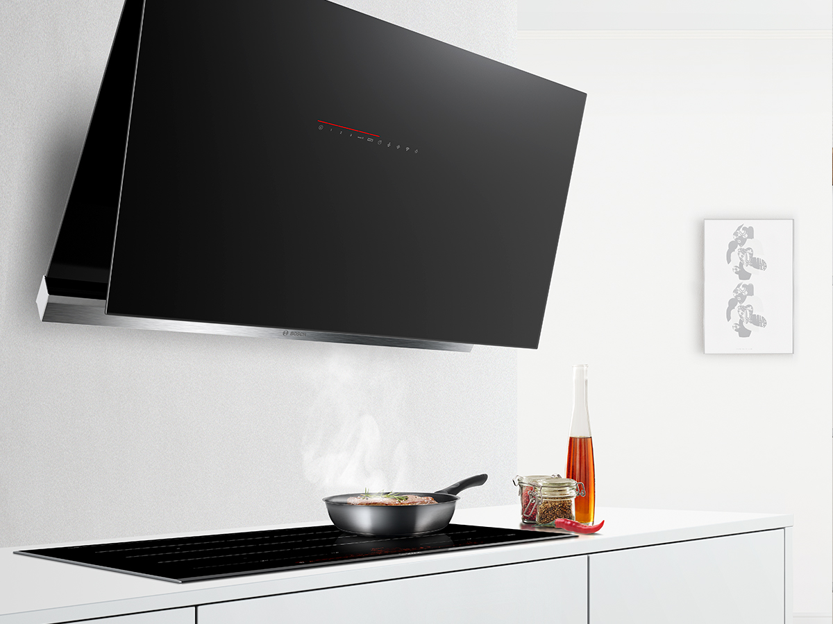 Cocina moderna con campana extractora negra sobre placa con sartén humeante. Botella, tarro y guindilla sobre encimera blanca. Cuadro en pared.