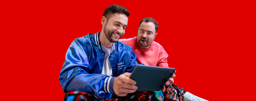 Dos hombres jóvenes sonríen mientras miran juntos una tablet sobre un fondo rojo.