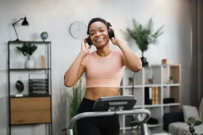 Mujer sonriente con audífonos en caminadora. Estantes blancos y negros y plantas en el fondo.