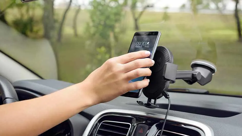 Una mano sostiene un teléfono móvil en un soporte negro dentro de un coche, con un parabrisas y vegetación visible al fondo.