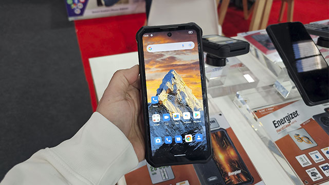 Mano sujetando un teléfono móvil negro con una montaña en la pantalla, sobre una mesa con folletos de Energizer.