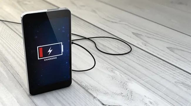 Un teléfono móvil negro muestra un icono de batería baja y un rayo, conectado a un cable sobre una superficie de madera.