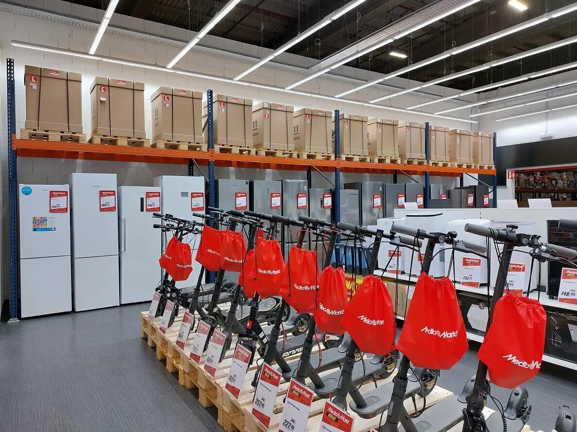 Interior de tienda con neveras blancas y patinetes negros con bolsas rojas. Cajas de cartón apiladas en estantes superiores.
