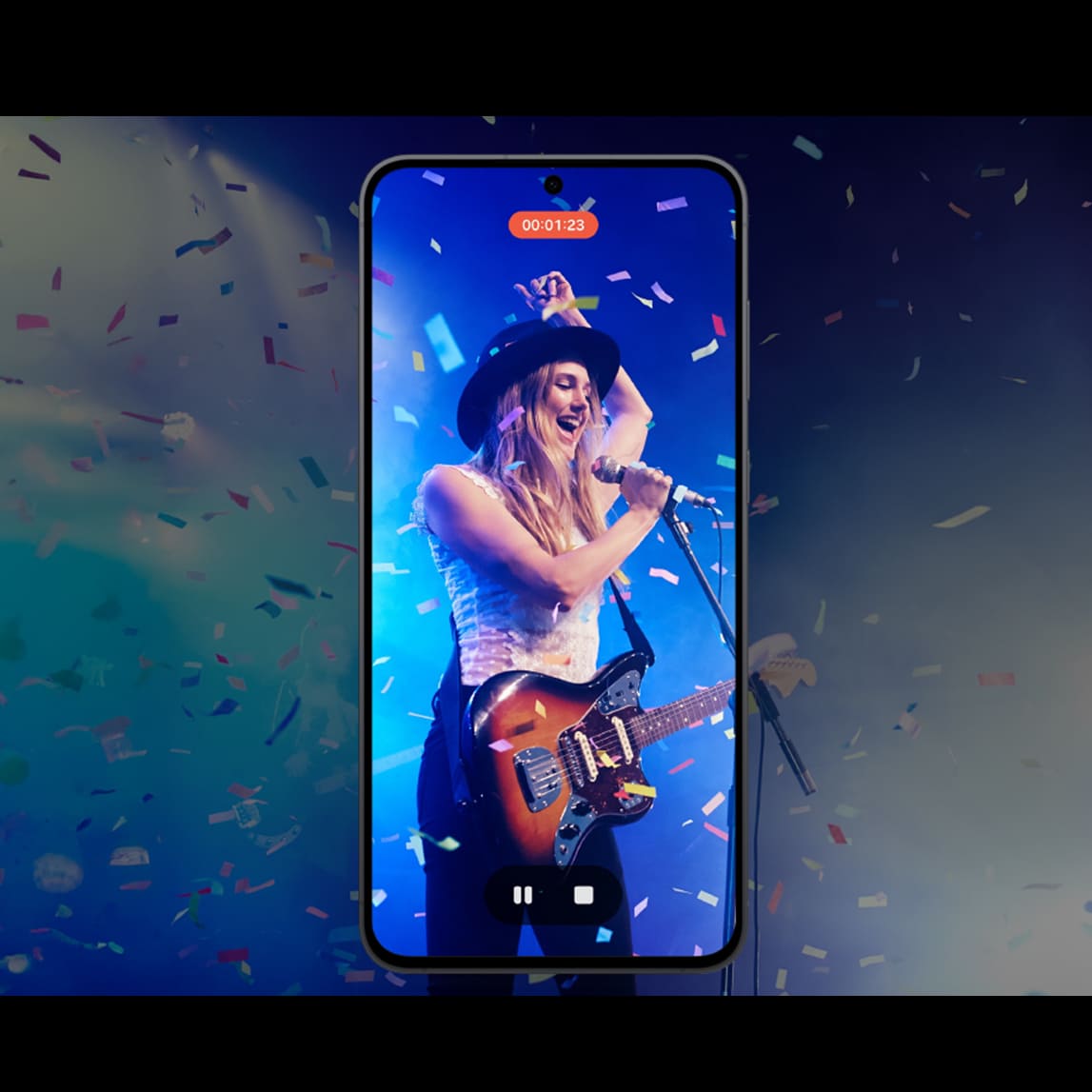 Un smartphone Samsung en position verticale enregistre une vidéo d'une femme jouant de la guitare électrique lors d'un concert, avec des lumières et des confettis en arrière-plan.