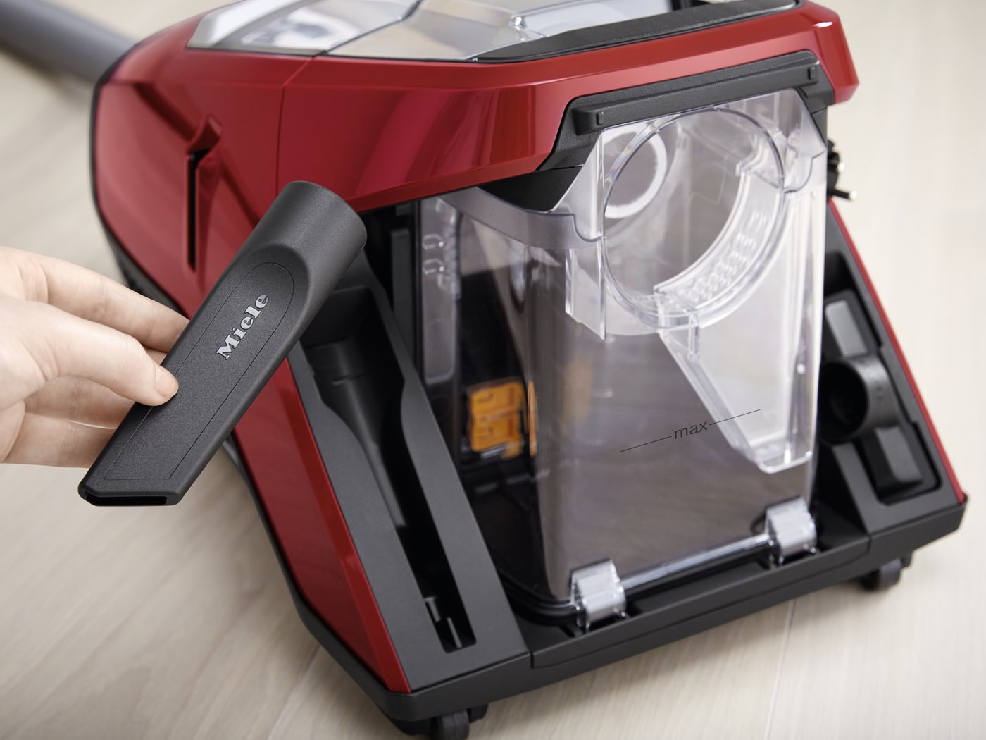 Aspiradora roja y negra con un depósito transparente donde una mano sostiene un accesorio negro con la marca "Miele".