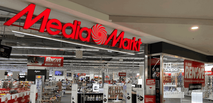 Entrada a tienda Media Markt con logo rojo y blanco. El interior tiene estanterías con electrónica y carteles promocionales.