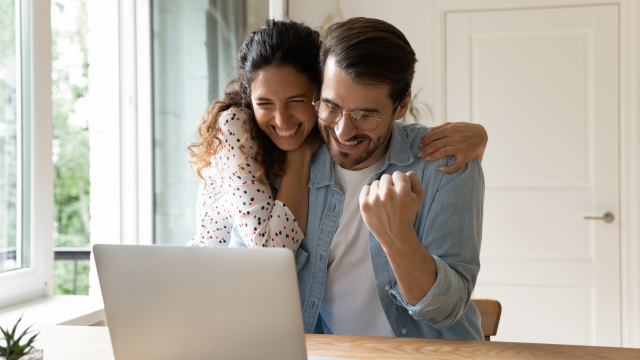 Una pareja sonriente frente a un portátil en una mesa de madera, celebrando con el puño cerrado. Ventana y puerta blancas al fondo.