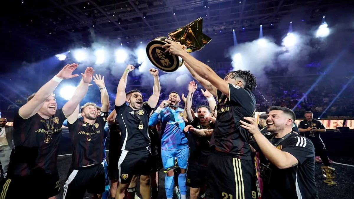 Jugadores de fútbol celebran con un trofeo dorado sobre un fondo oscuro y luces brillantes.