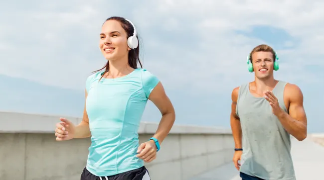 Una mujer y un hombre corren con auriculares. Ella lleva una camiseta cian y pantalones cortos negros. Él, una camiseta de tirantes gris.