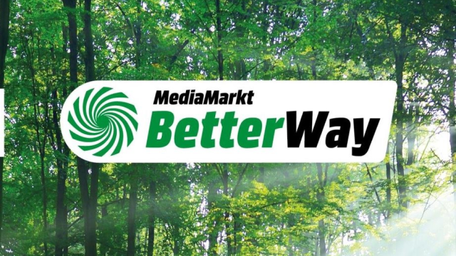 Un letrero blanco con "MediaMarkt BetterWay" en negro y verde, sobre un fondo de árboles verdes.