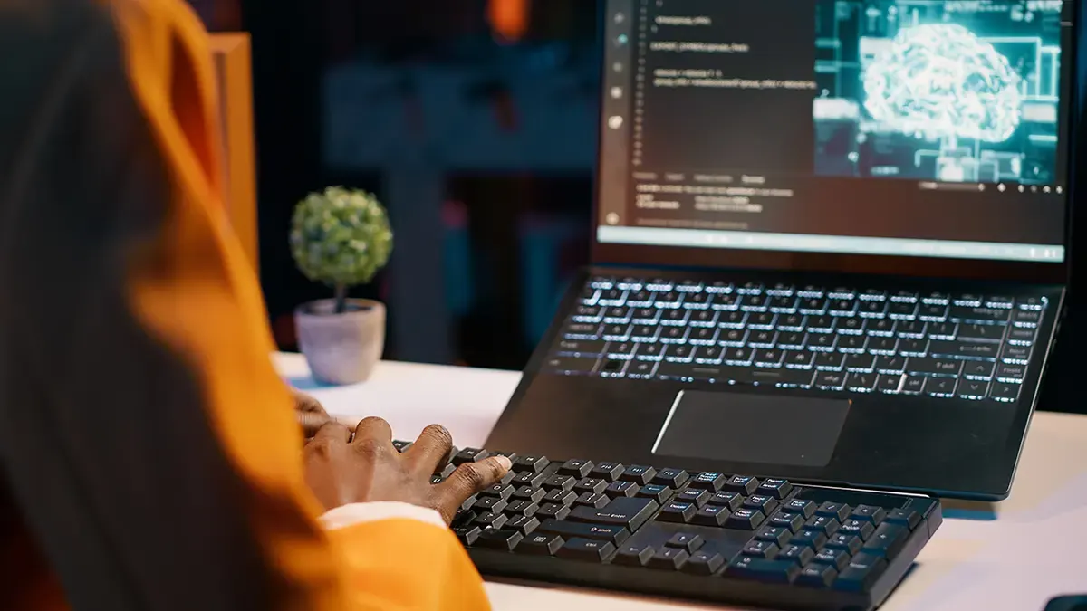 Persona tecleando en un teclado negro frente a una laptop. En la pantalla se ve código y una imagen de un cerebro. Hay una planta pequeña.