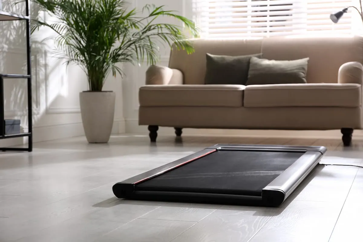 Cinta de correr compacta plegable en el suelo de un salón moderno, frente a un sofá beige y una planta grande.