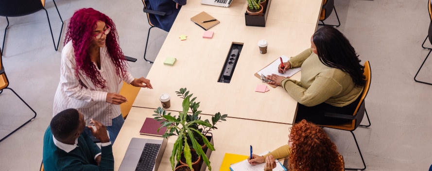 Vista superior de cuatro profesionales trabajando en equipo alrededor de mesa blanca con computadoras, papeles y planta como decoración