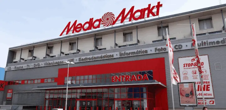 Edificio de Media Markt con letrero rojo en la parte superior y la palabra "Entrada" sobre la puerta. Banderas rojas y blancas ondean cerca.