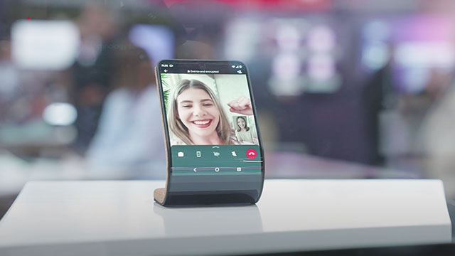 Un teléfono flexible está sobre una mesa blanca, mostrando una videollamada con dos personas. El fondo es borroso.