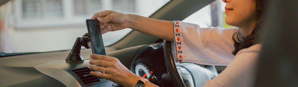 Mujer colocando un móvil negro en un soporte en el salpicadero de un coche. Lleva reloj y una camisa blanca con bordados.