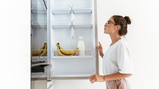 Mujer abre nevera blanca con plátanos y botella. La nevera está casi vacía y la mujer lleva una camisa blanca.