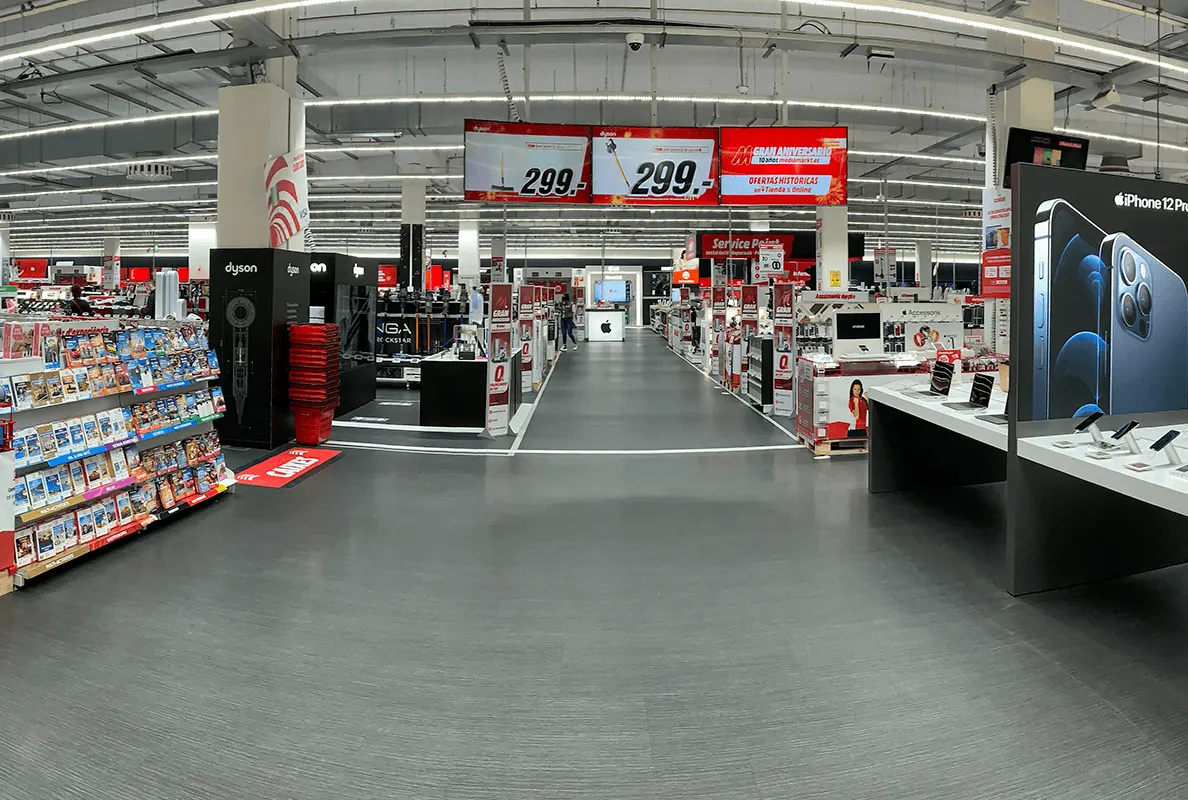 Pasillo de tienda con estanterías de productos a los lados. Señalización digital suspendida y mesas de exhibición al frente.