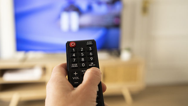 Mano sujetando un mando a distancia negro con botones numéricos frente a un televisor encendido y un mueble de madera.