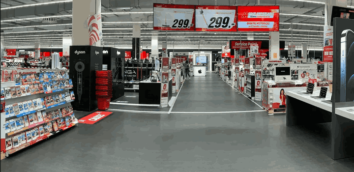 Pasillo de tienda con estanterías de productos electrónicos, carteles rojos y blancos, y suelo gris.