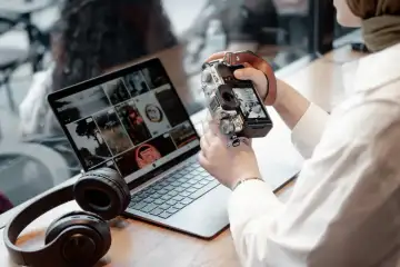 Persona revisando una cámara junto a un portátil con auriculares sobre la mesa.