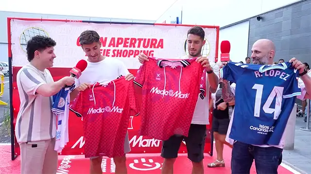 Cuatro hombres muestran camisetas rojas y azules con números frente a un cartel rojo y blanco en un evento al aire libre.