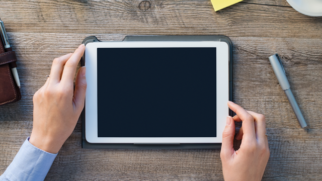 Vista superior de manos sosteniendo una tableta blanca con pantalla oscura sobre una mesa de madera con bolígrafo y accesorios.