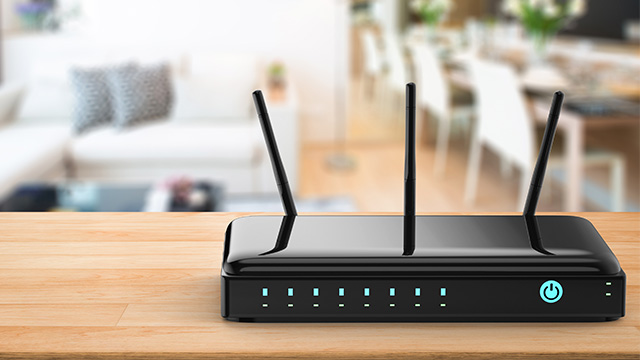 Un router negro con tres antenas se encuentra sobre una mesa de madera. En el fondo, se ve una sala de estar desenfocada.