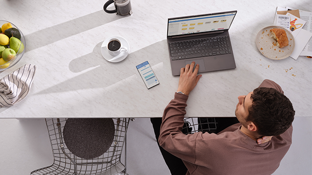Vista superior de hombre trabajando en laptop sobre mesa blanca con fruta, café y plato. Un smartphone está a su lado.
