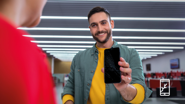 Un hombre sonriente muestra un móvil con la pantalla rota. Lleva una camisa verde sobre una camiseta amarilla, delante de luces blancas.