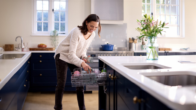 Mujer sonriente vaciando un lavavajillas en una cocina luminosa. Hay flores en un jarrón, fregaderos dobles y electrodomésticos modernos.