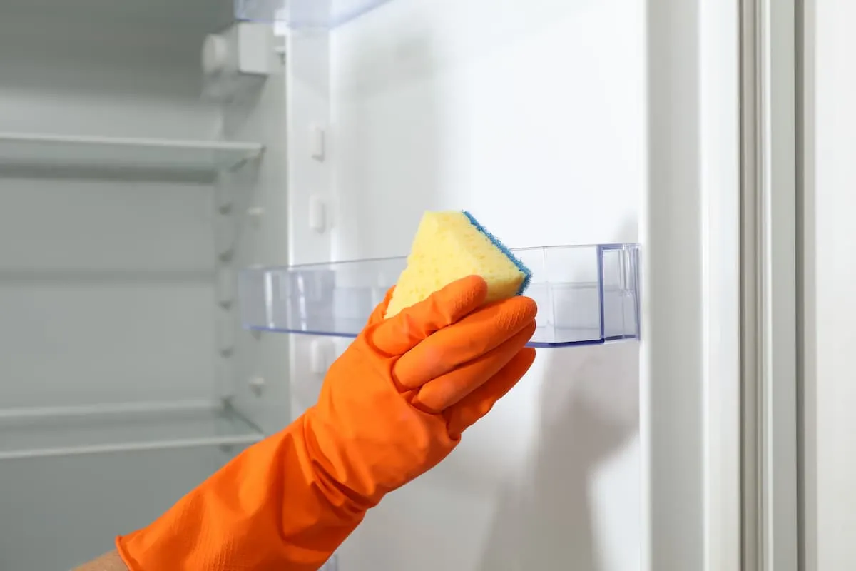 Mano con guante naranja limpiando el estante de la puerta del refrigerador con una esponja