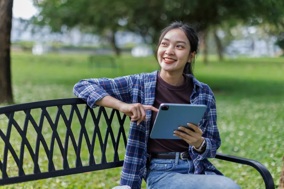 Mujer joven sonriente sentada en un banco de un parque, sosteniendo una tablet.
