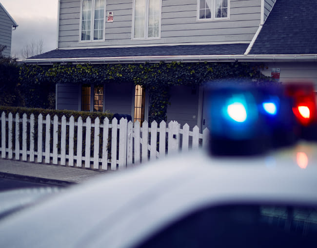 Casa unifamiliar gris con valla blanca y luces interiores encendidas al atardecer, con coche policial desenfocado en primer plano y luces azules rojas activadas.
